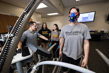 Person wearing a mask is walking on a device while people in the back wearing ASU shirts look at screens