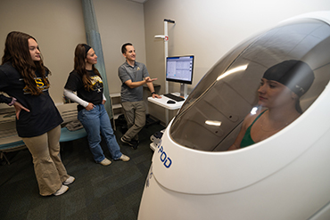 Person in a bodpod container wearing a swim cap while three people look at a screen