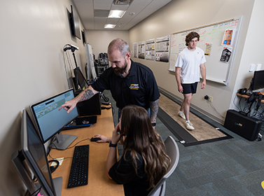 Skinner shows a student the screen while a person walks behind them on a gait mat
