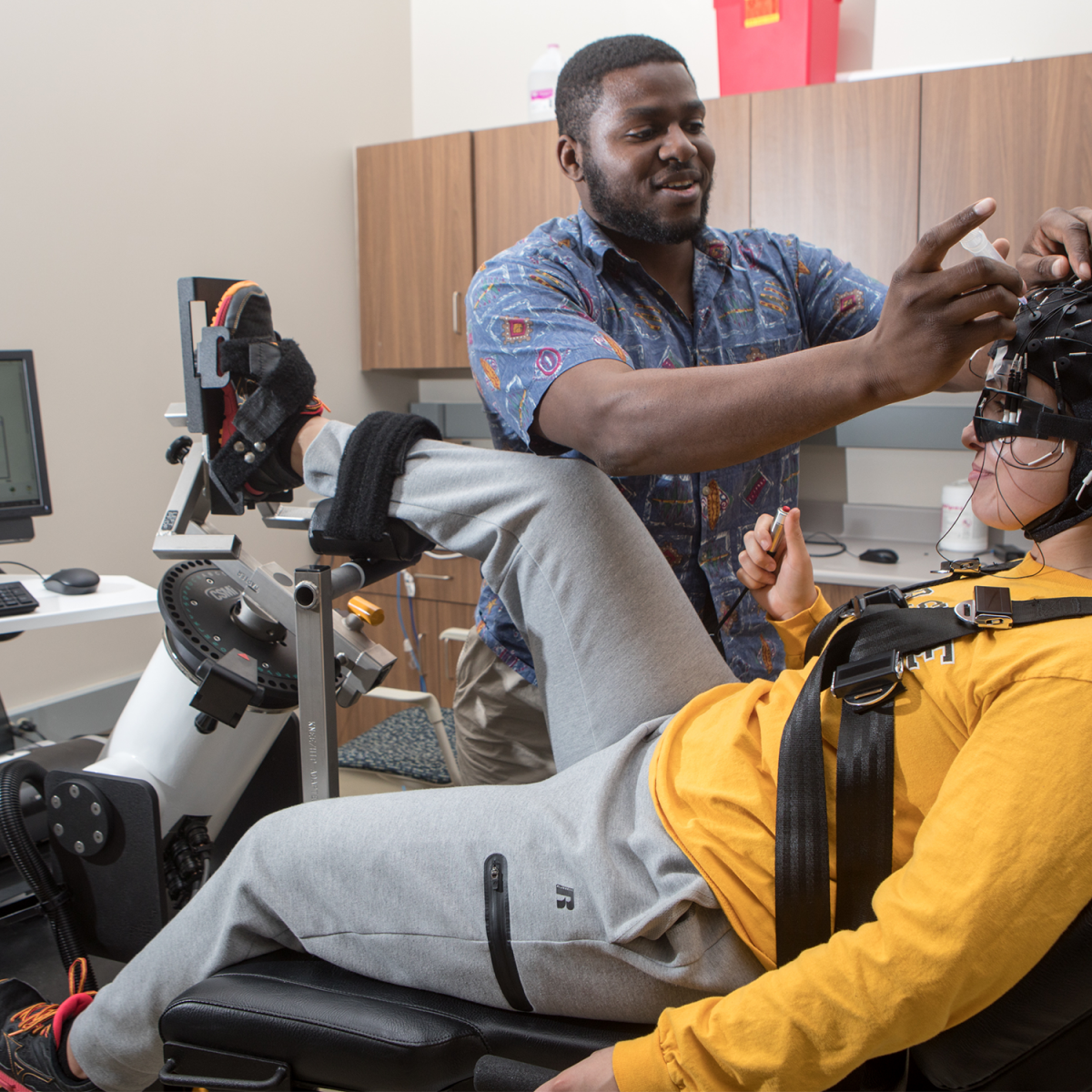 A student researcher adjusting an electroencephalography cap on someone sitting on a dynamometer